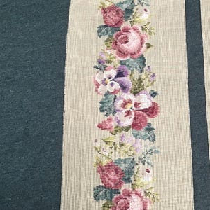 May include: Needlepoint canvas with a floral design featuring pink roses, purple pansies, and green leaves. The canvas is beige and has a vertical floral pattern. A bag of colourful yarn is visible.
