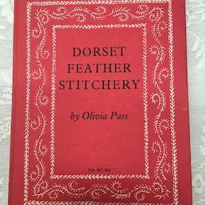 May include: A vintage book titled "Dorset Feather Stitchery" by Olivia Pass. The book has a red cover with white decorative borders and text. The cover also includes the price "10s. 6d. net".