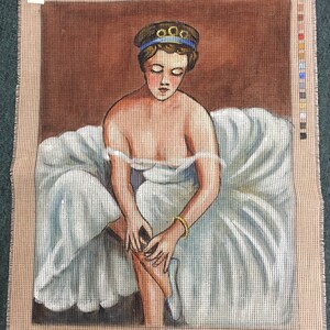 May include: A needlepoint canvas depicting a woman in a white dress and blue headband, sitting on a brown background. She is holding a white shoe in her hands and appears to be adjusting it. The canvas is labeled "99-515" and "181/7" in the top right and bottom right corners, respectively.