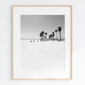 May include: Black and white photograph of a beach with palm trees and people riding bicycles. The beach is empty and the sky is clear.