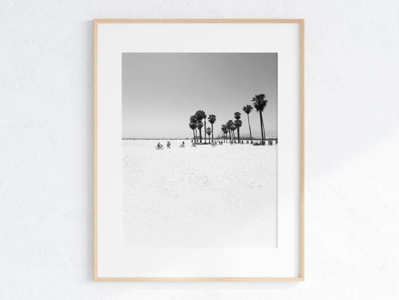 May include: Black and white photograph of a beach with palm trees and people riding bicycles. The beach is empty and the sky is clear.