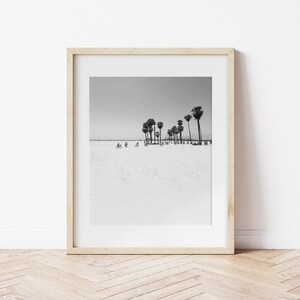 May include: Black and white photograph of palm trees on a beach with people riding bicycles in the distance. The photo is framed in a light wood frame.