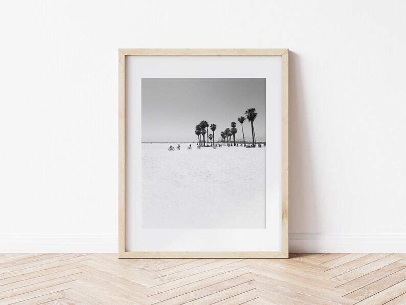 May include: Black and white photograph of palm trees on a beach with people riding bicycles in the distance. The photo is framed in a light wood frame.