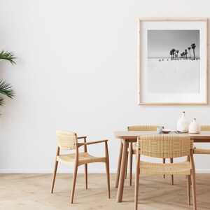 May include: A dining room with a wooden table and six woven chairs. A framed black and white photograph of palm trees on a beach hangs on the wall. A potted plant is in the corner of the room.