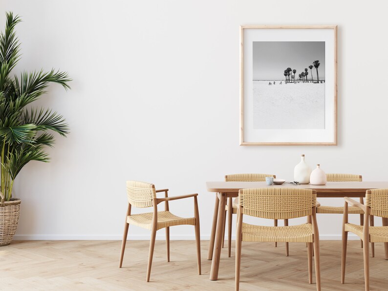 May include: A dining room with a wooden table and six woven chairs. A framed black and white photograph of palm trees on a beach hangs on the wall. A potted plant is in the corner of the room.