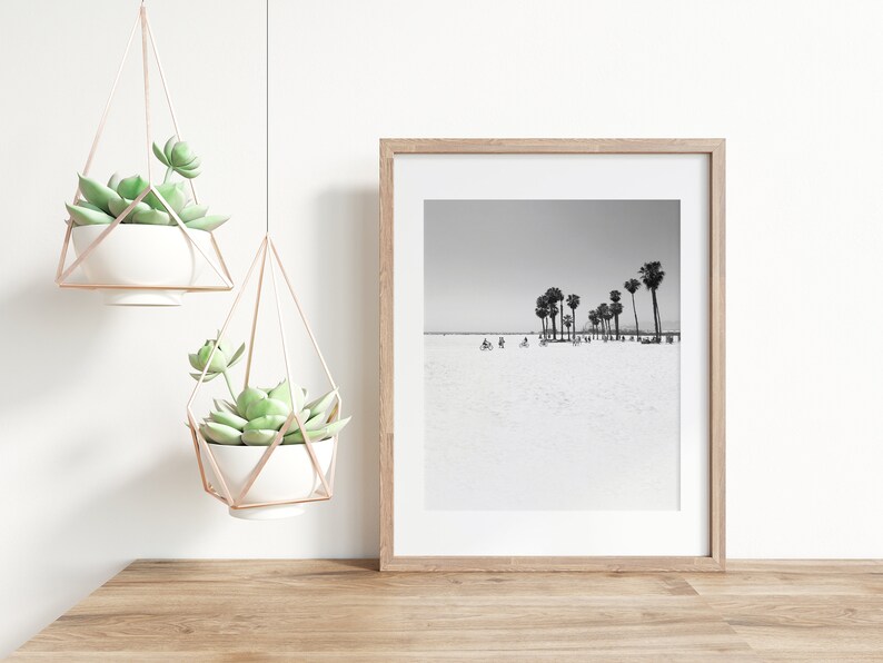 May include: A black and white photograph of palm trees on a beach. The photo is framed in a light wood frame and hanging on a white wall.