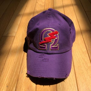 May include: A purple baseball cap with a distressed look. The cap has a gold and red embroidered patch with a lightning bolt and the letter omega.