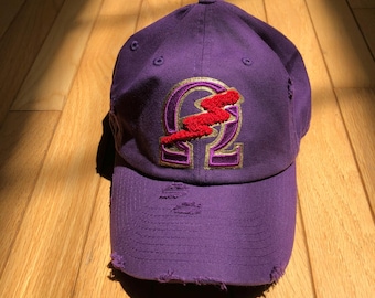 Könnte beinhalten: Eine lilafarbene Baseballkappe mit einem abgenutzten Look. Die Kappe hat einen goldenen und roten gestickten Aufnäher mit einem Blitz und dem Buchstaben Omega.