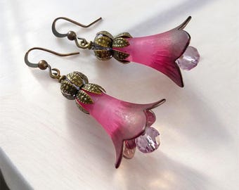 May include: A pair of drop earrings featuring pink, bell-shaped flower petals made of a translucent material. Each earring is topped with an antique bronze-toned metal cap and finished with a small, faceted, light purple crystal bead at the base.