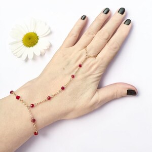 May include: A gold-toned hand chain featuring a delicate metal link chain adorned with small, faceted red beads. The jewellery piece connects a bracelet section to a single ring component, creating a decorative accessory for the hand.