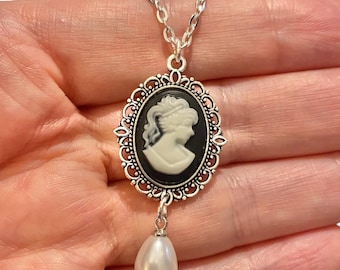 May include: Silver-toned necklace featuring an oval cameo pendant with a profile of a woman in white against a black background. The pendant has a decorative filigree border and a dangling pear-shaped pearl.