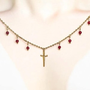 Könnte beinhalten: Eine goldfarbene Halskette mit einem kleinen Dolch-Anhänger und mehreren hängenden roten Perlen. Die Halskette ist filigran und scheint aus Kettengliedern gefertigt zu sein. Die Perlen sind tiefrot und verleihen dem Design einen Farbtupfer.