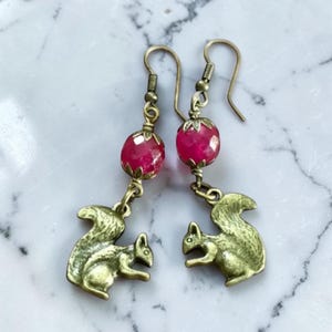 Könnte beinhalten: Ein Paar Ohrhänger mit eichhörnchenförmigen Anhängern in Antikmessing-Optik, die unter facettierten, runden, pinkfarbenen Perlen hängen. Die Perlen sind mit kleinen metallenen Kappen verziert und an einfachen Ohrhaken aus Messing befestigt.