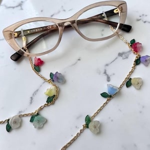 May include: A pair of translucent beige cat-eye glasses with the brand name Vogue printed on the temple, attached to a gold-tone metal chain featuring small, multi-coloured floral charms in shades of pink, purple, blue, yellow, and white.