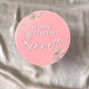 May include: Pink round cake topper with silver glitter accents. The topper says "Happy Birthday Bennett" in white lettering.