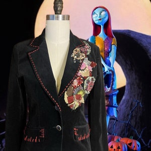 May include: Black velvet blazer with red stitching and decorative patches. The blazer has a single-button closure and two front pockets. The patches are made of various fabrics and embellishments. The blazer is displayed on a mannequin.