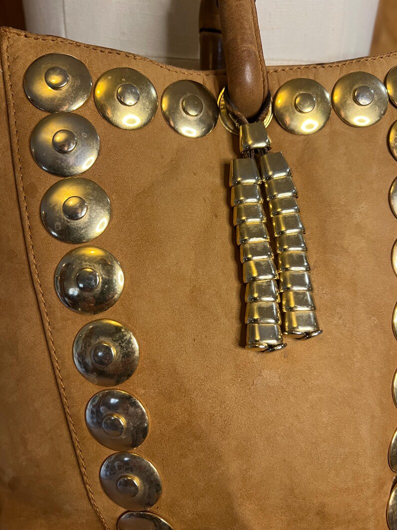 May include: A brown leather tote bag with gold metal accents and a tassel. The bag has a row of round metal studs down the side.