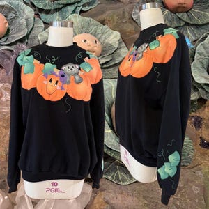 May include: Black sweatshirt featuring a Halloween-themed design. The design includes three orange pumpkins with smiling faces and a gray teddy bear. Green leaves and vines accent the pumpkins. The sweatshirt is displayed on a mannequin.