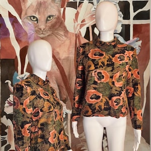 May include: Two mannequins showcase a long-sleeved top with a floral design. The top has a high collar and a repeating pattern of orange and black flowers on a green and brown backdrop. A large painting of a cat is in the background.