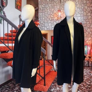 May include: Two mannequins display a black coat with a wide collar and long sleeves. The coat is knee-length and has a simple, classic design. The mannequins are positioned in front of a staircase with orange carpet and a stone wall.