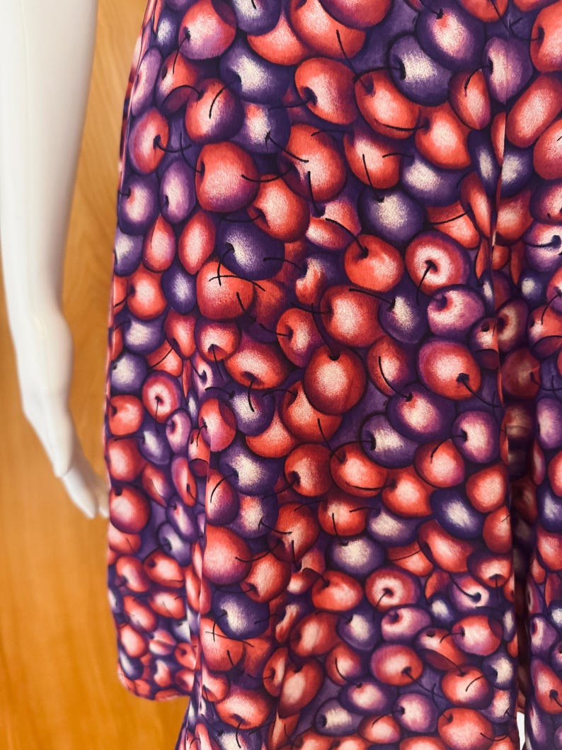 Peut inclure: Un v&ecirc;tement &agrave; motif de cerises. Le tissu pr&eacute;sente un motif r&eacute;p&eacute;t&eacute; de cerises rouges, violettes et blanches. Les cerises ont une apparence r&eacute;aliste avec des reflets et des ombres, cr&eacute;ant un aspect textur&eacute;. Le v&ecirc;tement semble &ecirc;tre une jupe ou une robe.