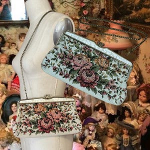 May include: Two vintage floral tapestry handbags. One is off-white with a pink and peach rose pattern, the other is light blue with a similar floral design. Both have metal frames and chain straps, showcasing a classic, elegant style.