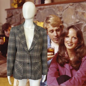 May include: A gray plaid blazer with a two-button closure and two front pockets. The blazer is displayed on a mannequin. The background features a retro-style image of people.