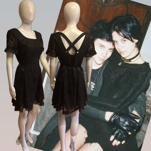 May include: Two mannequins display a black dress with a floral pattern. The dress features short sleeves, a scoop neckline, and a flared skirt. The back of the dress has a crisscross design. A vintage photograph is in the background.