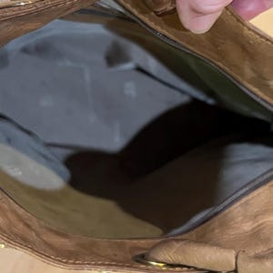 May include: A brown leather tote bag with a zipper closure and a gold metal zipper pull. The bag is open and the inside is lined with a light brown fabric.