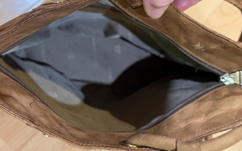 May include: A brown leather tote bag with a zipper closure and a gold metal zipper pull. The bag is open and the inside is lined with a light brown fabric.