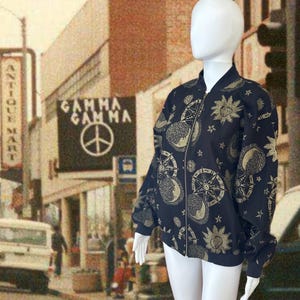 May include: A navy blue bomber jacket with a gold celestial pattern featuring suns, moons, stars, and compass designs. The jacket has a zip-up front and ribbed cuffs. The background shows a vintage street scene with a sign that reads "Gamma Gamma".