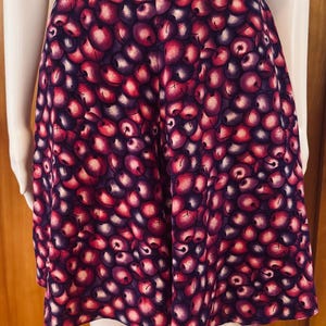 Peut inclure: Jupe trap&egrave;ze avec un motif de cerises vives. La jupe pr&eacute;sente une taille haute et une silhouette &eacute;vas&eacute;e, avec un motif r&eacute;p&eacute;t&eacute; de cerises rouges et violettes sur un fond violet fonc&eacute;. La jupe est pr&eacute;sent&eacute;e sur un mannequin.