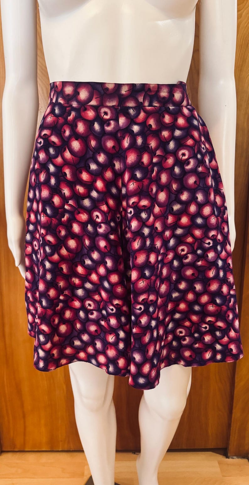 Peut inclure: Jupe trap&egrave;ze avec un motif de cerises vives. La jupe pr&eacute;sente une taille haute et une silhouette &eacute;vas&eacute;e, avec un motif r&eacute;p&eacute;t&eacute; de cerises rouges et violettes sur un fond violet fonc&eacute;. La jupe est pr&eacute;sent&eacute;e sur un mannequin.