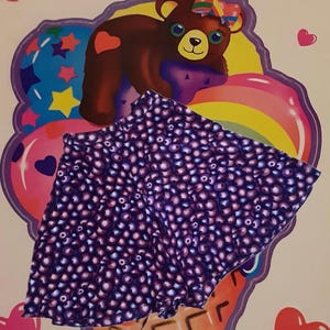 Peut inclure: Un short violet orn&eacute; d'un motif de petits cercles violets clairs. Le short est pos&eacute; sur un fond color&eacute; repr&eacute;sentant un ours de dessin anim&eacute;, un arc-en-ciel et des c&oelig;urs. Le short semble &ecirc;tre fait d'un tissu doux.