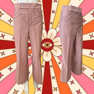 May include: Dusty rose-colored pants with a high waist and flared leg. The pants feature two front pockets and a button closure. The stitching is a darker shade of pink. The pants are displayed on a mannequin against a retro-style background.