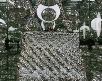 May include: A silver-toned handbag featuring a metallic lattice overlay with embedded faceted beads. The bag has a structured frame, a top clasp closure, and a matching silver chain handle. The bottom edge is finished with a row of dangling beads.