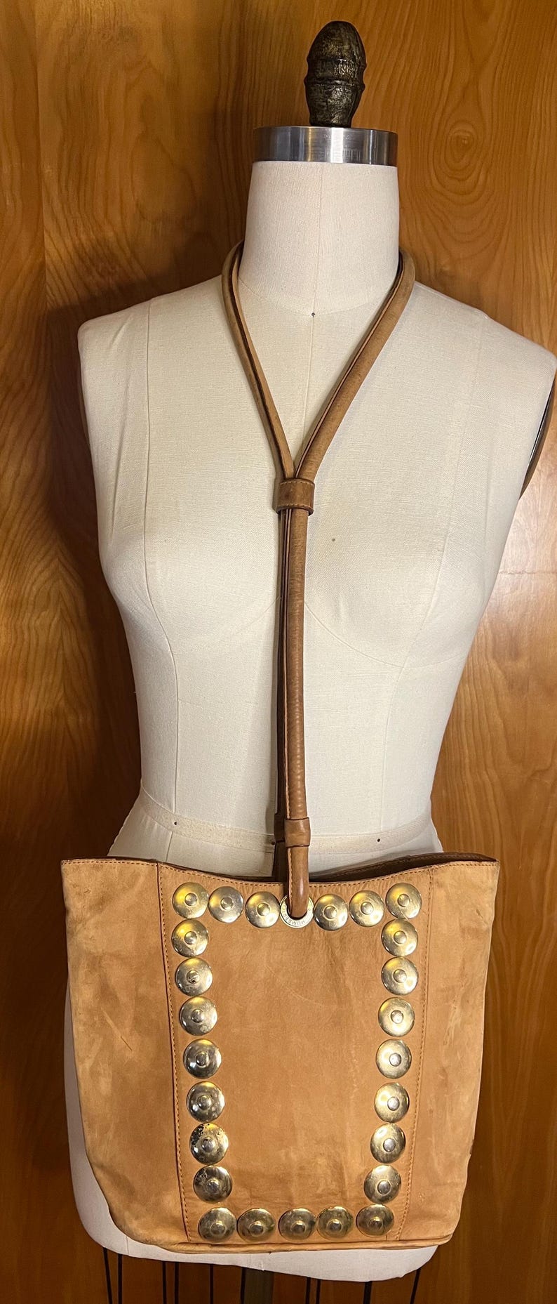 May include: A tan suede tote bag with a long tan leather strap. The bag has a decorative border of silver metal circles.