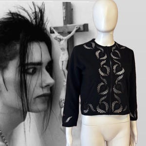 May include: Black cardigan with intricate openwork design on a mannequin. The jacket features a round neckline, long sleeves, and a zipper closure. The openwork pattern is a repeating floral design. The background features a black and white image of a person and a crucifix.