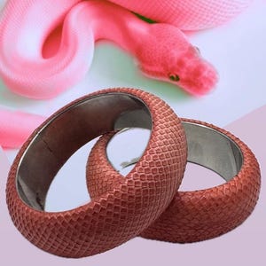 May include: Two wide bangle bracelets featuring a textured, reddish-brown exterior with a pattern resembling snake scales and a smooth, metallic silver-toned interior lining. The bracelets are positioned in front of a bright pink snake.