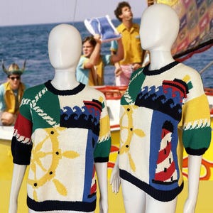 Könnte beinhalten: Ein bunter, kurzärmeliger Strickpullover mit nautischem Thema. Der Pullover hat ein Patchwork-Design mit einem Leuchtturm, einem Steuerrad und Wellen in Rot, Blau, Grün, Gelb und Weiß.