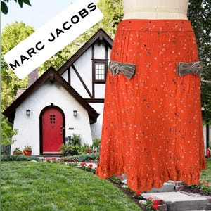 May include: An orange skirt with a floral pattern and two grey velvet bow accents. The skirt has a ruffled hem and is displayed on a mannequin. The text "MARC JACOBS" is in a white rectangle in the background.