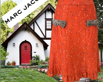 Peut inclure: Jupe orange à motif floral avec deux nœuds en velours gris. La jupe a un ourlet à volants et est présentée sur un mannequin. Le texte "MARC JACOBS" est dans un rectangle blanc en arrière-plan.