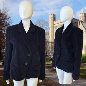 Pode incluir: Um blazer de veludo cotelê preto com lapela entalhada, dois botões frontais e dois bolsos com aba. A jaqueta tem mangas compridas com punhos dobrados e um design de ombro estruturado, exibido em dois manequins brancos.