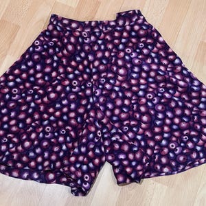 Peut inclure: Un short violet avec un motif de petites formes rondes, violet fonc&eacute; et roses. Le short est fait d'un tissu l&eacute;ger et a une jambe large et &eacute;vas&eacute;e. Il est pos&eacute; sur une surface en bois brun clair.