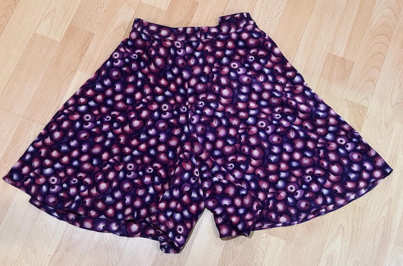 Peut inclure: Un short violet avec un motif de petites formes rondes, violet fonc&eacute; et roses. Le short est fait d'un tissu l&eacute;ger et a une jambe large et &eacute;vas&eacute;e. Il est pos&eacute; sur une surface en bois brun clair.