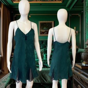 May include: A dark green satin slip dress with a bow detail at the neckline. The dress has a fitted bodice and a flowing skirt with a scalloped hem.
