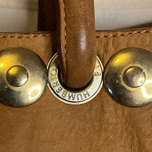 May include: Close-up of a brown leather handbag with gold-toned hardware. The hardware features the text "HUMBERT" and a small "F" on the side.