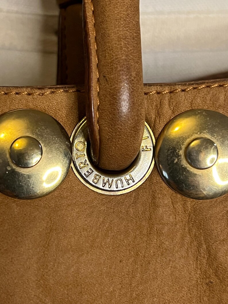 May include: Close-up of a brown leather handbag with gold-toned hardware. The hardware features the text "HUMBERT" and a small "F" on the side.