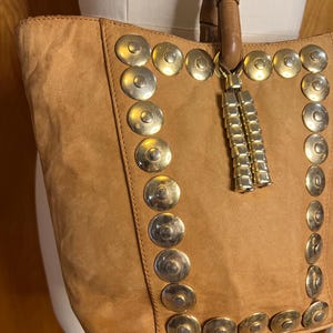 May include: A brown suede tote bag with gold metal accents. The bag has a long tassel hanging from the top.