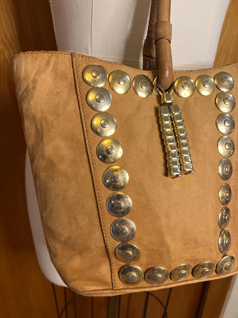 May include: A brown suede tote bag with gold metal accents. The bag has a long tassel hanging from the top.